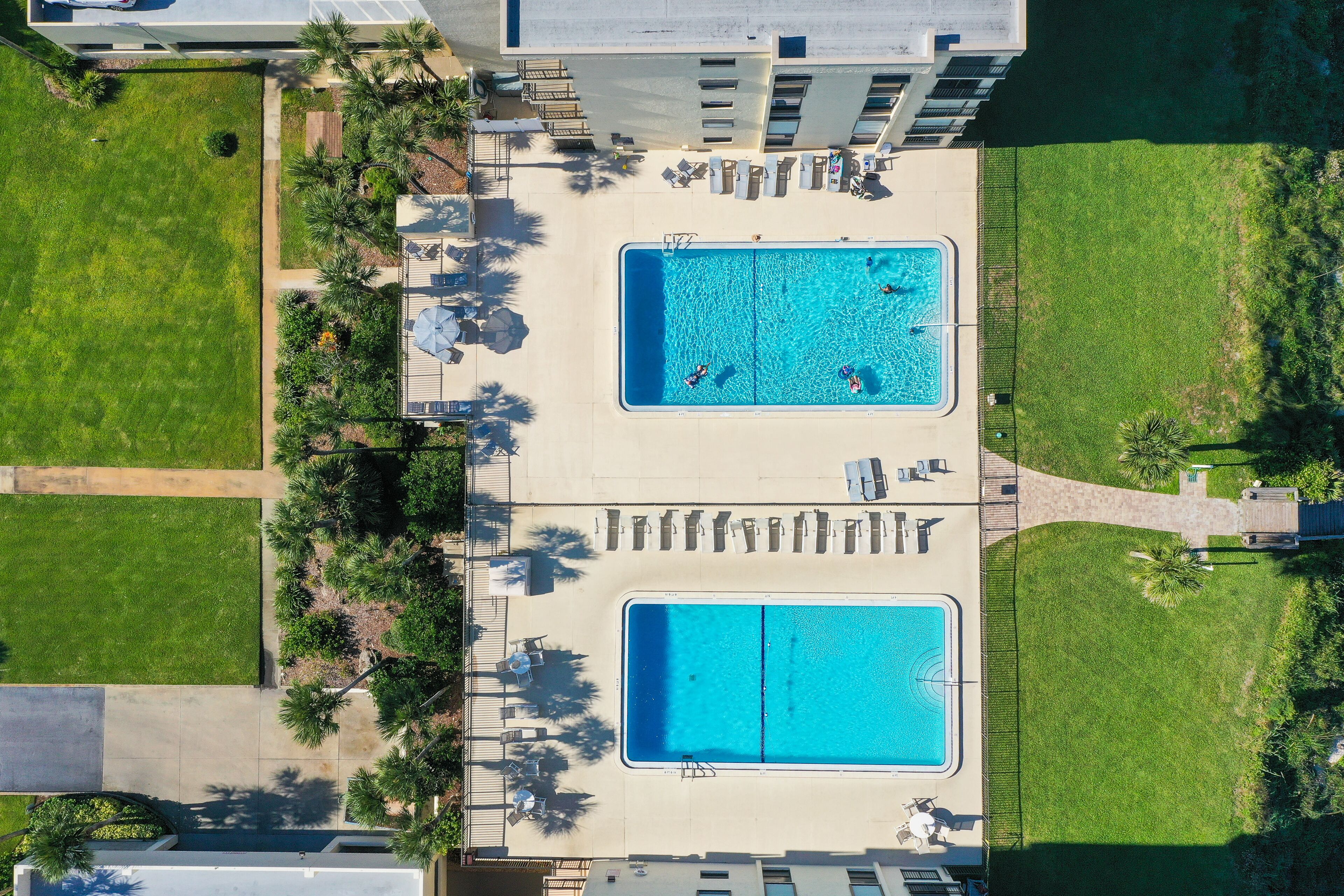 An aerial view of a hotel with two adjacent swimming pools, surrounded by lounge chairs and palm trees.