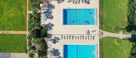 An aerial view of a hotel with two adjacent swimming pools, surrounded by lounge chairs and palm trees.