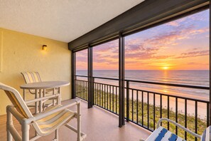A balcony with a table and chairs, overlooking a beach at sunset.
