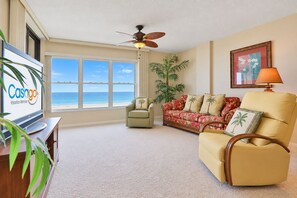 A spacious living room with a large window, a ceiling fan, a television, and a sofa with tropical-themed cushions.