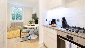Une cuisine moderne avec des meubles blancs, une cuisinière à gaz, une table à manger avec des chaises et une fenêtre donnant sur des arbres.