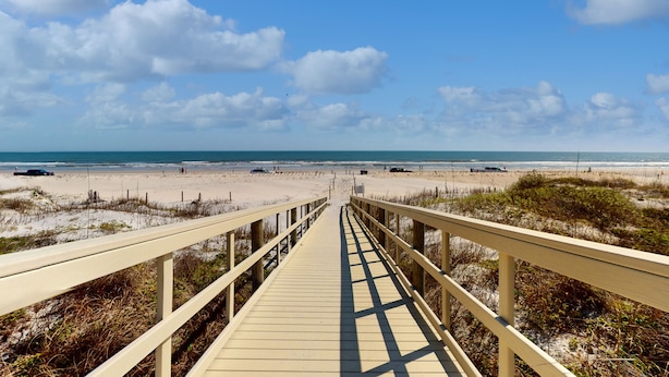 A wooden boardwalk leading to a beach with a clear sky and distant ocean view.