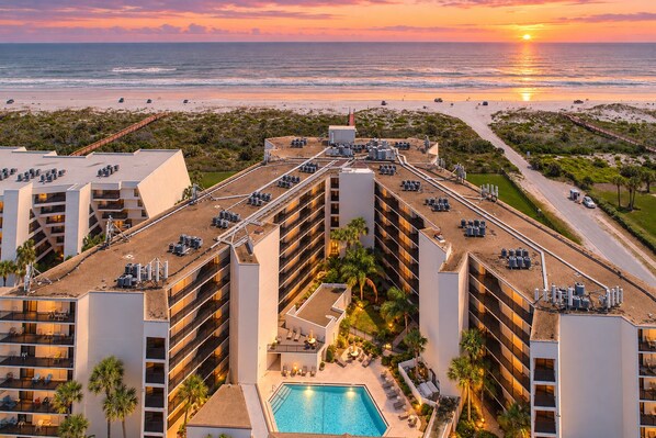 A hotel with multiple floors, a rooftop pool, and a view of the beach and ocean at sunset.