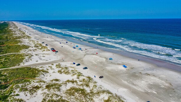 Une vue aérienne d’une plage avec une longue étendue de sable, quelques parasols et quelques voitures garées le long du rivage.