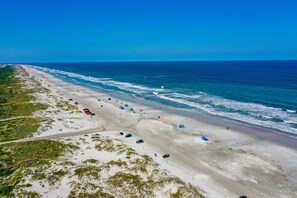 An aerial view of a beach with a long stretch of sand, a few umbrellas, and a few cars parked along the shore.