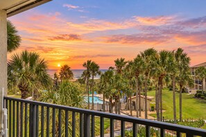 A balcony with a view of a sunset over the ocean, palm trees, and a swimming pool.