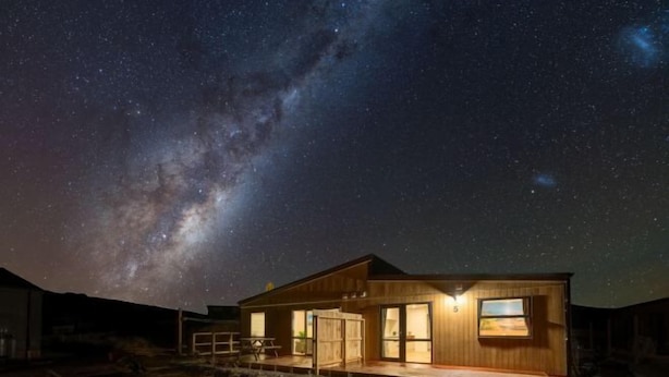 Una cabaña de madera con un amplio ventanal, un patio y vistas de la Vía Láctea.