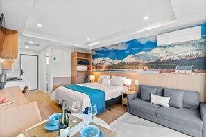 A hotel room with a bed, a sofa, a dining area with a table and chairs, and a large painting of a mountain landscape. - Sunshine Suites (Lake Tekapo)
