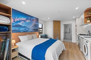 Una habitación de hotel moderna con una cama grande, un cuadro de un paisaje montañoso, una lavadora y una kitchenette.