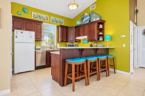 A kitchen with wooden cabinets, a refrigerator, a microwave, and a bar area with stools. - Two-level home with partial ocean view, balcony, WiFi, & W/D - walk to the beach (St. Augustine)