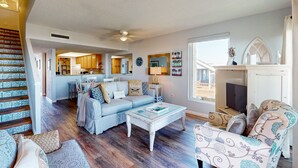 Amplia sala de estar con un sofá azul claro, una mesa de centro blanca y un sillón estampado. Una escalera con barandilla decorada conduce a una zona de cocina.