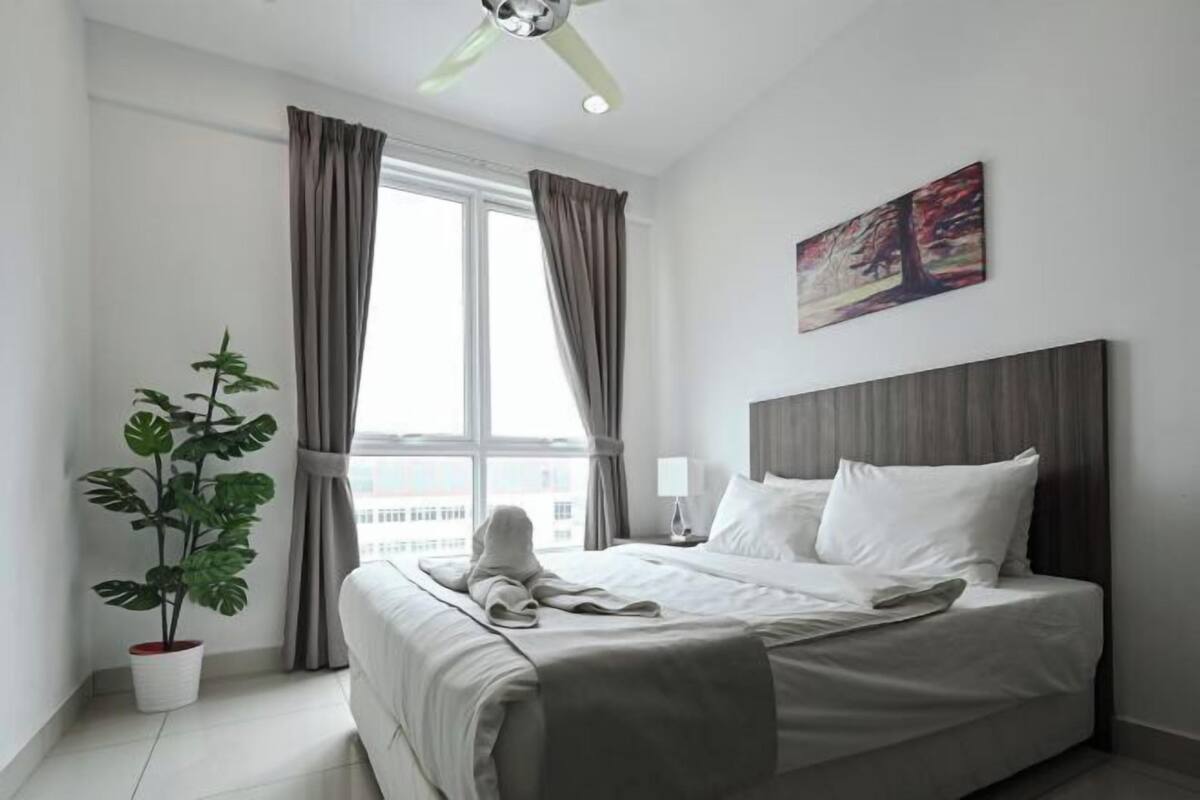 A hotel room with a bed, a potted plant, a window with curtains, a painting on the wall, and a ceiling fan.