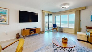 A modern living room with a flat-screen TV, wooden furniture, a balcony with a view of the beach, and a large window. - Oceanfront condo with pool, ocean views, and washer/dryer (St. Augustine)