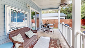 A covered porch with a wicker sofa, a dining table with chairs, and a large window with a view of a house and trees. - Charming home steps from the beach with enclosed yard and grill - dog-friendly (St. Augustine)