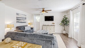 A living room with a grey sofa, a wooden coffee table with a tray of cookies, a ceiling fan, a television, and a potted plant.
