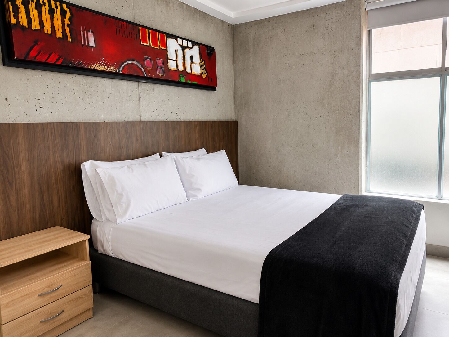 A modern bedroom with a bed, a wooden nightstand, a large abstract painting, and a window.