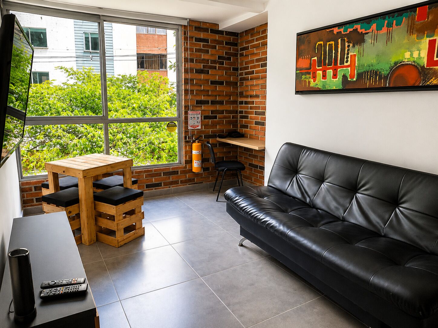 A modern living room with a black leather sofa, wooden table, and chairs, a large window with a view of greenery, and a fire extinguisher on the wall.
