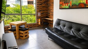 A modern living room with a black leather sofa, wooden table, and chairs, a large window with a view of greenery, and a fire extinguisher on the wall. - Best bang for your buck in Laureles (Medellín)