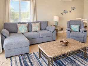 A living room with a grey sectional sofa, a matching armchair, a small table with a lamp, and decorative wall art.
