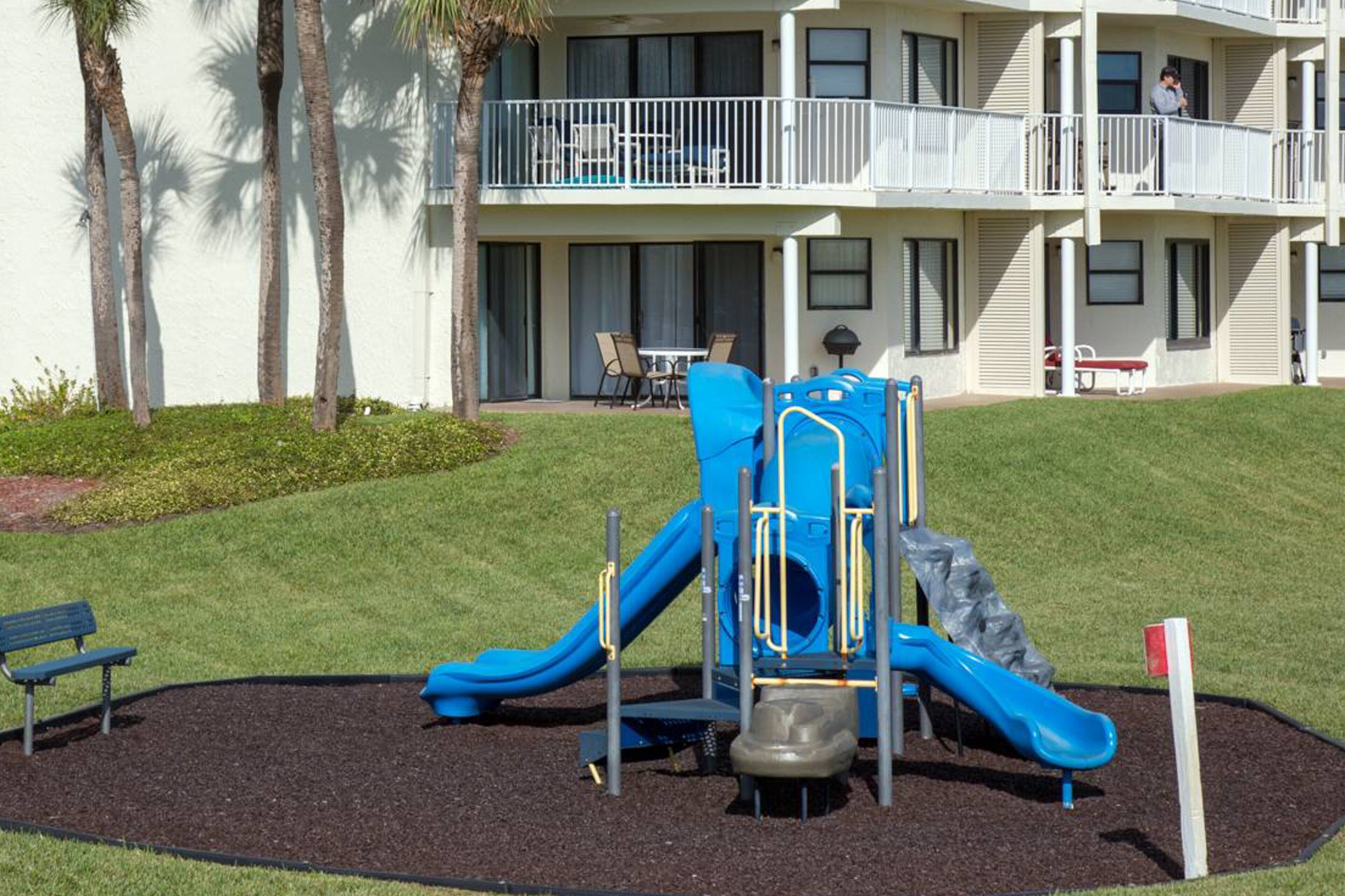 A playground with a blue slide and climbing structure, surrounded by a landscaped area with a bench and palm trees.