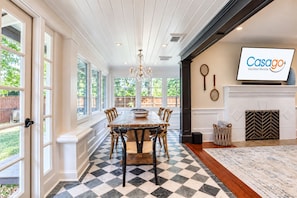 A dining area with a checkered floor, a wooden table, and chairs. There is a fireplace with a TV on the wall.