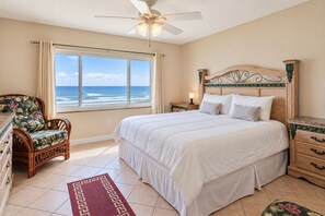 A bedroom with a bed, a chair, a nightstand, a dresser, a window with a view of the ocean, and a ceiling fan.