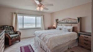 Quarto com cama, cadeira, mesa de cabeceira, ventilador de teto e janela com cortinas.