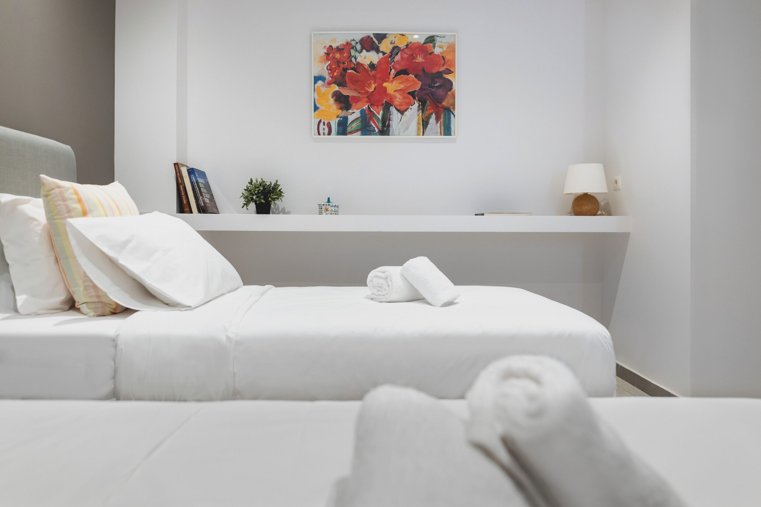 Ein modernes Hotelzimmer mit einem ordentlich bezogenen Bett, einer kleinen Ablagefläche mit Büchern und einer Pflanze sowie einem farbenfrohen Bild an der Wand.
