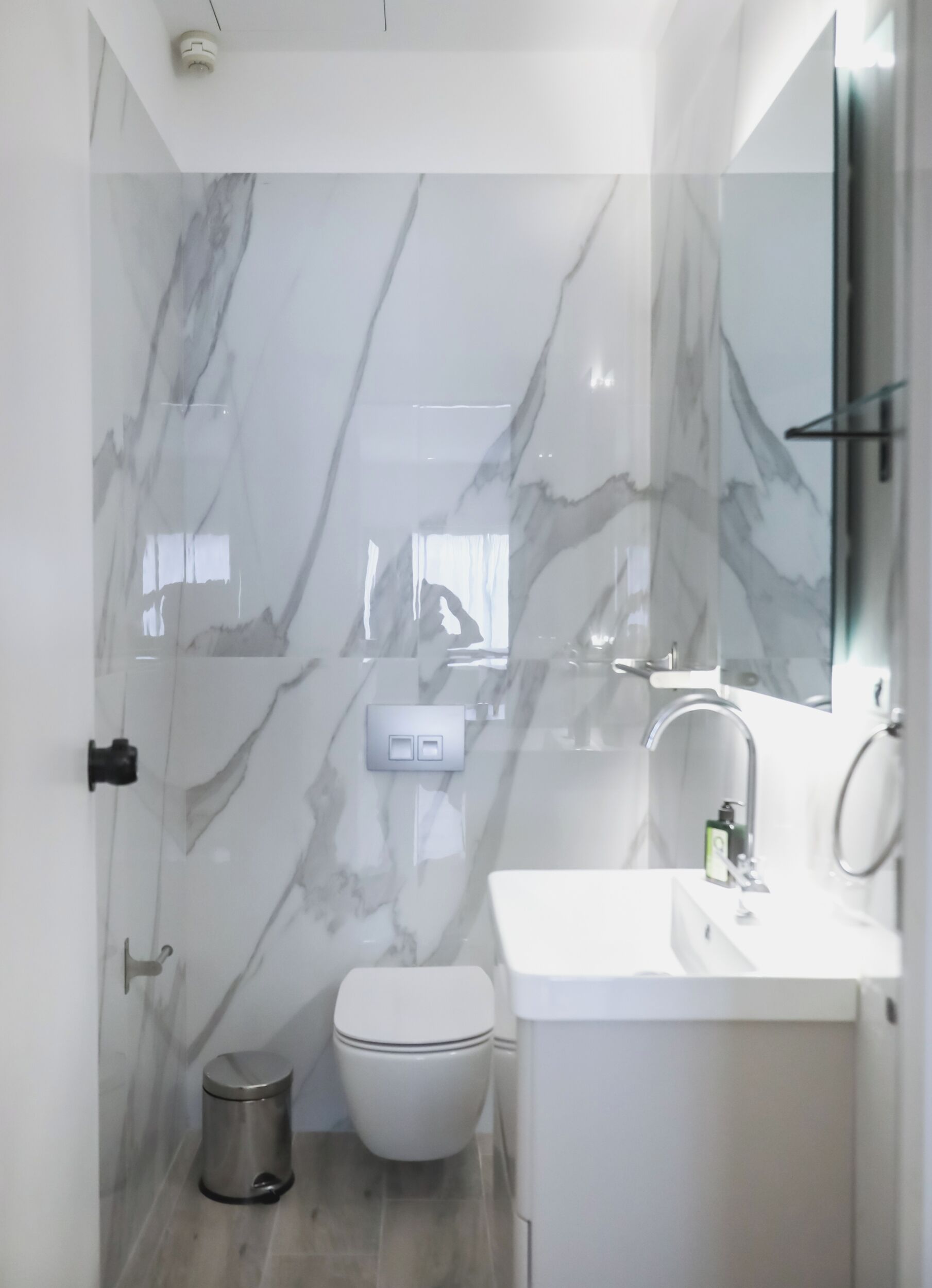 A modern bathroom with a marble wall, a toilet, a sink, and a trash bin.