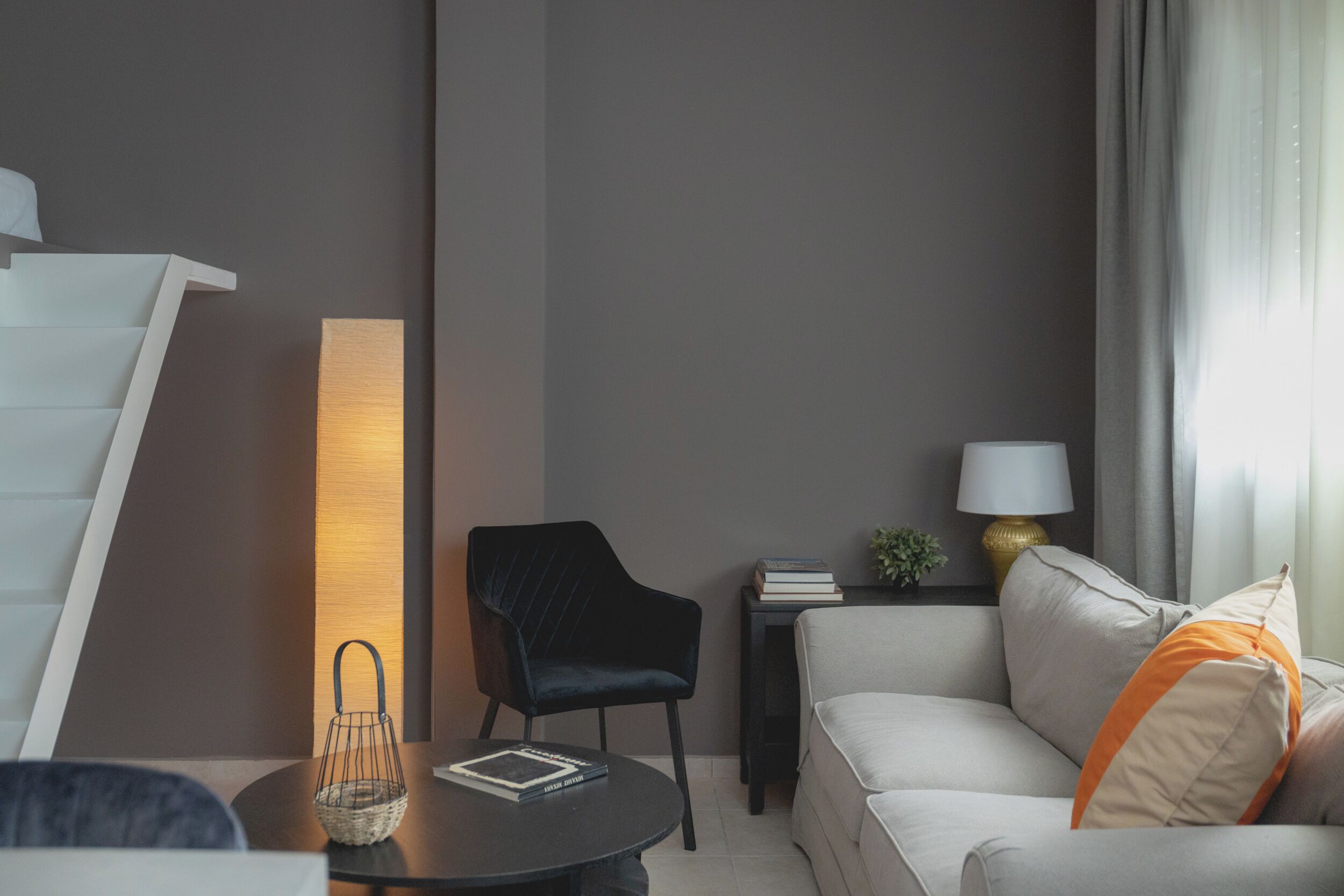 A modern living room with a sofa, a black chair, a small table with a lamp, and a staircase.