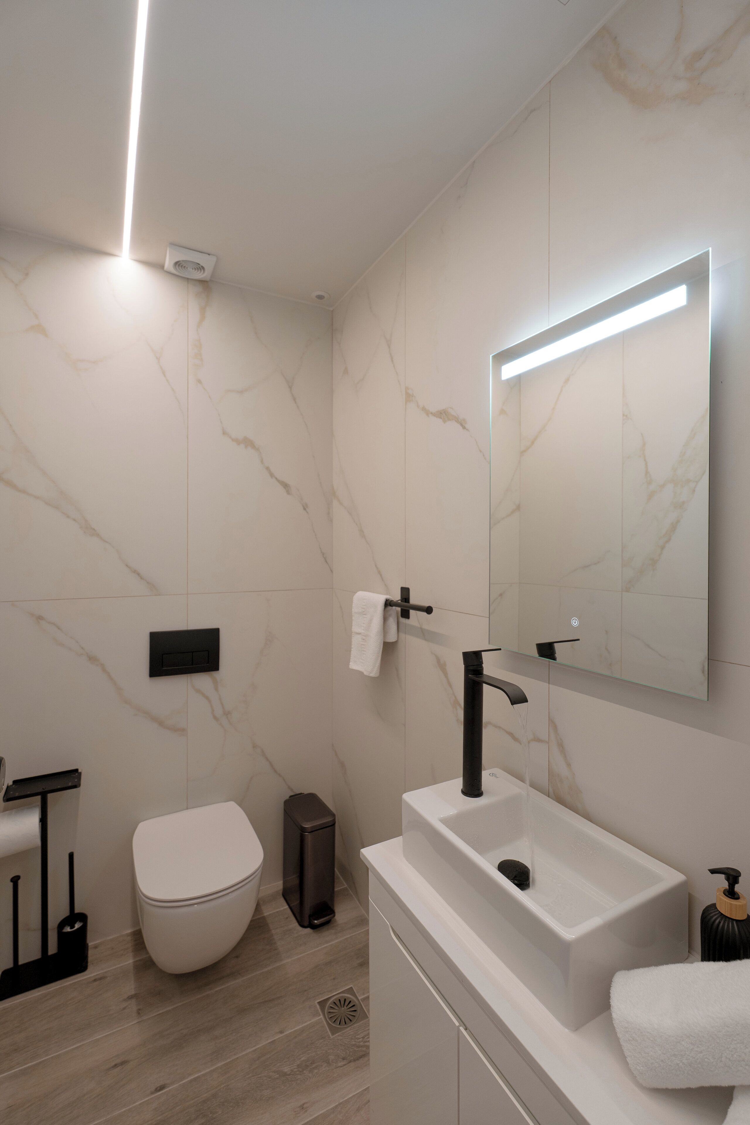 A modern bathroom with a white marble wall, a rectangular sink with a black faucet, a white toilet, and a mirror with integrated lighting.