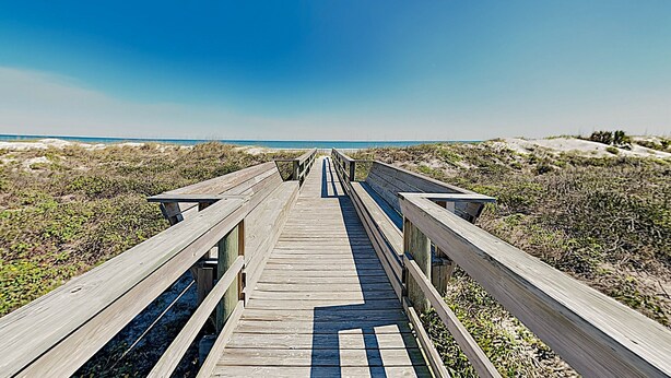 Une promenade en bois qui mène à une plage bordée de dunes et dotée d’une végétation clairsemée.