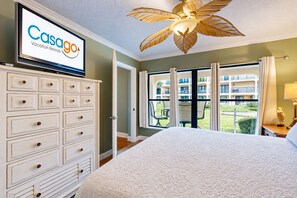 A hotel room with a bed, a dresser, a television displaying 'Casago Vacation Rentals,' and a view of a building and greenery outside.