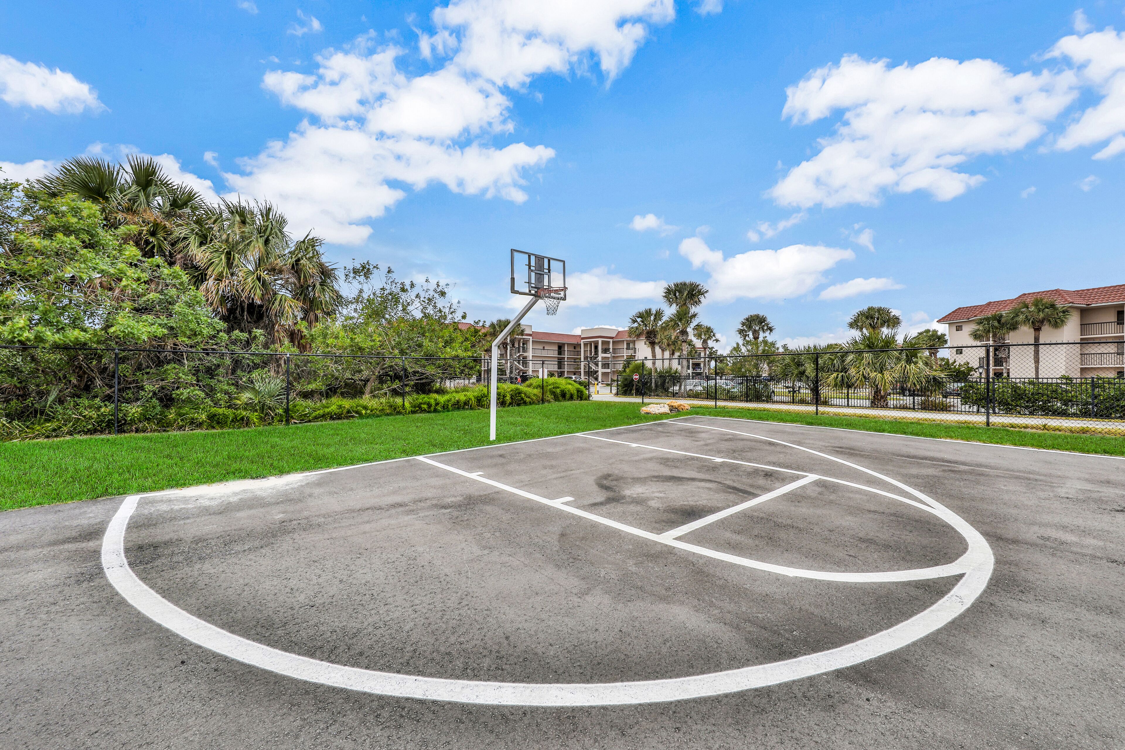 An outdoor basketball court with a net, surrounded by greenery and buildings.