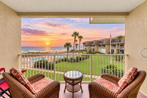 A balcony with wicker furniture, a glass table, and a view of the beach and ocean at sunset.