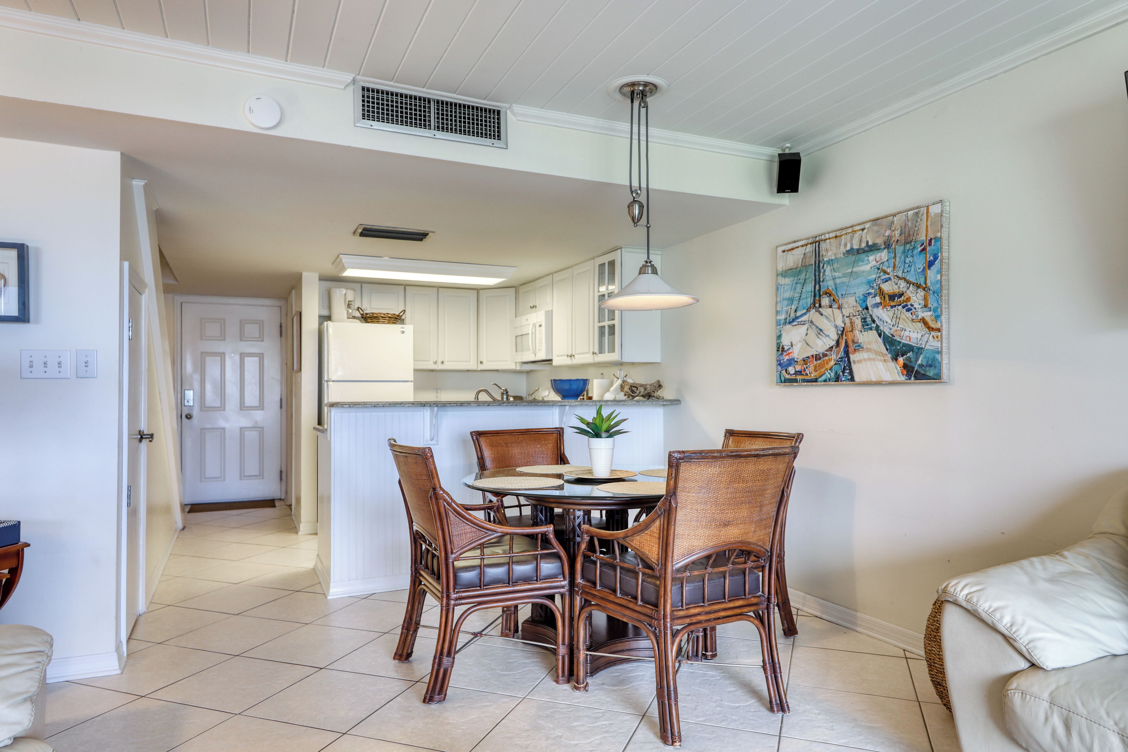 A dining area with a round table and four chairs, a kitchen in the background, and a painting on the wall.
