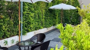 Un patio al aire libre con una mesa de vidrio y sillas de mimbre, rodeado de abundante vegetación y un paraguas blanco.