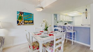 A dining area with a white table and chairs, a hanging light fixture, and a kitchen area with white cabinets and a blue countertop. - Oceanview condo with beach access, pool, tennis courts and private patio (St. Augustine)