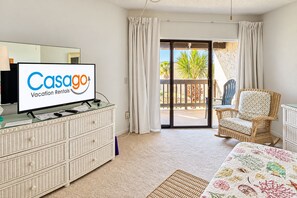 A hotel room with a television displaying 'Casago Vacation Rentals,' a wicker dresser, a wicker chair, a bed with a floral bedspread, and a sliding glass door leading to a balcony with a view of palm trees.