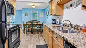 A kitchen with wooden cabinets, a granite countertop, a sink, and a dining area with chairs and a table. - Charming Condo w/ Pool – 500 Yards to Beach, Near Lighthouse & Downtown (St. Augustine)