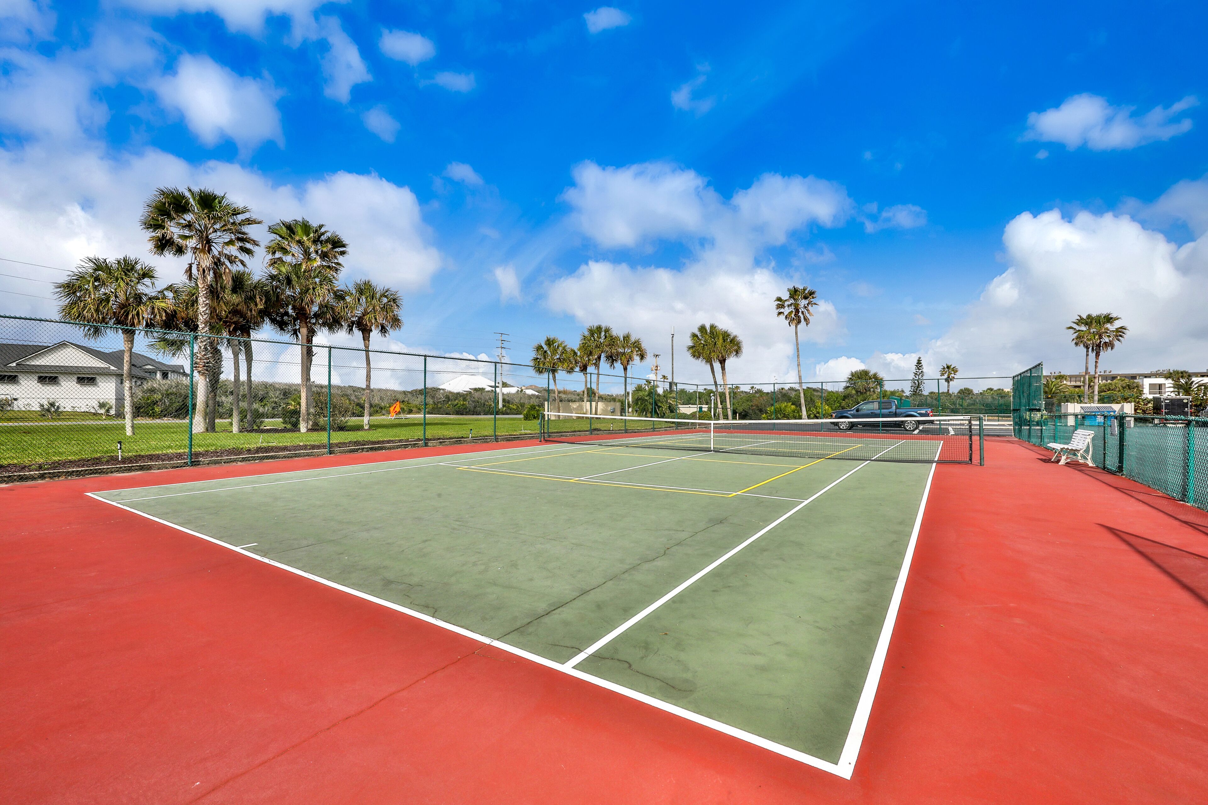 An outdoor tennis court with a red and green surface, surrounded by palm trees and a clear blue sky.