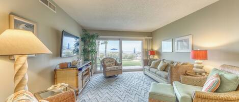 A spacious living room with a large window, wicker furniture, and a herringbone-patterned rug.
