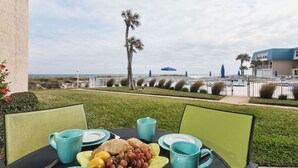 An outdoor dining area with a table set for two, featuring a plate of fruit, a pitcher, and cups. The setting includes a view of the beach and a palm tree, with a building and umbrellas in the background. - Island-themed living with furnished patio, shared pool & beach access (St. Augustine)