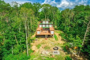A two-story cabin surrounded by dense forest, with a deck and outdoor seating area.