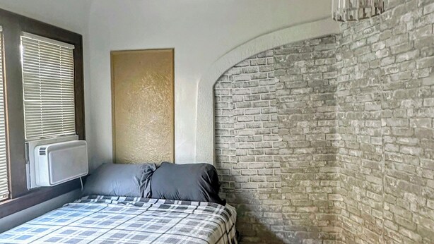 A bedroom with a bed, a wall-mounted air conditioner, a window with blinds, a chandelier, and a brick-patterned wall. - Step into this spacious-style home (Milwaukee)