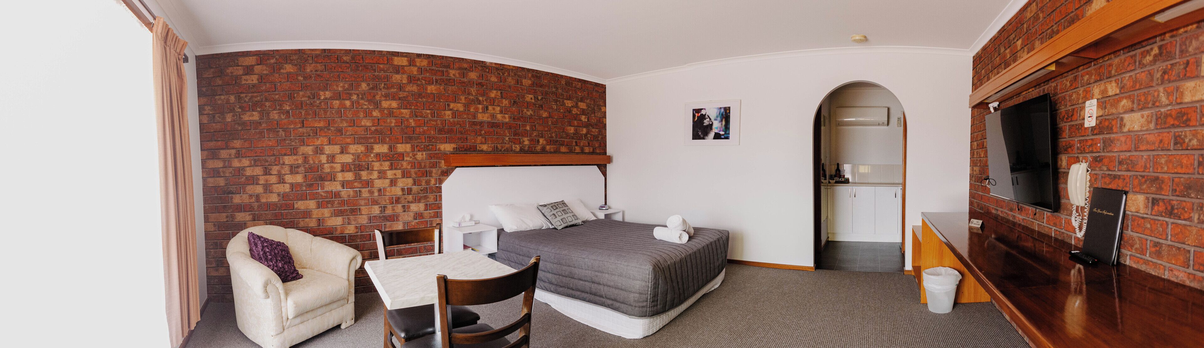A hotel room with a brick wall, a bed, a desk, a chair, a TV, and a bathroom.