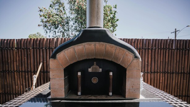 Un horno de ladrillo al aire libre con chimenea, sobre una base de baldosas, con un fondo de valla de bambú.