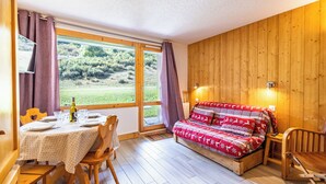 Una habitación acogedora con suelo de madera, una cama con manta estampada, una mesa de comedor preparada para dos personas y un amplio ventanal con vistas a zonas verdes.