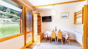 Une pièce chaleureuse avec un décor intérieur en bois, une télévision, une table avec des chaises et une vue sur un paysage verdoyant par une grande fenêtre.