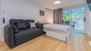 Una habitación moderna con un sofá negro, una cama con cabecera blanca y un ventanal corredero que da a un espacio al aire libre.